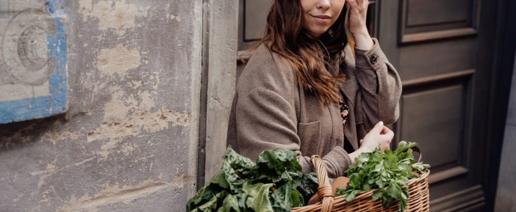 A woman smiling while holding a wicker basket filled with fresh green vegetables outdoors.