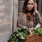 A woman smiling while holding a wicker basket filled with fresh green vegetables outdoors.