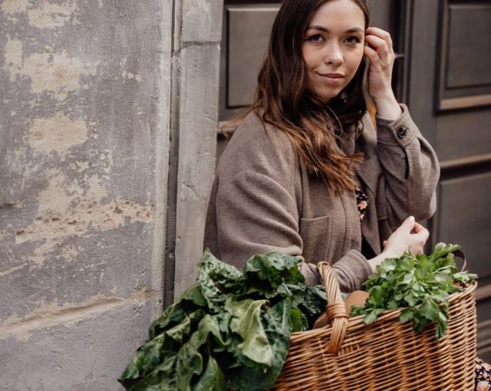 A woman smiling while holding a wicker basket filled with fresh green vegetables outdoors.