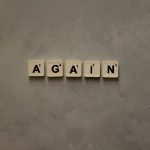 Close-up of Scrabble tiles spelling out 'Again' on a textured background.